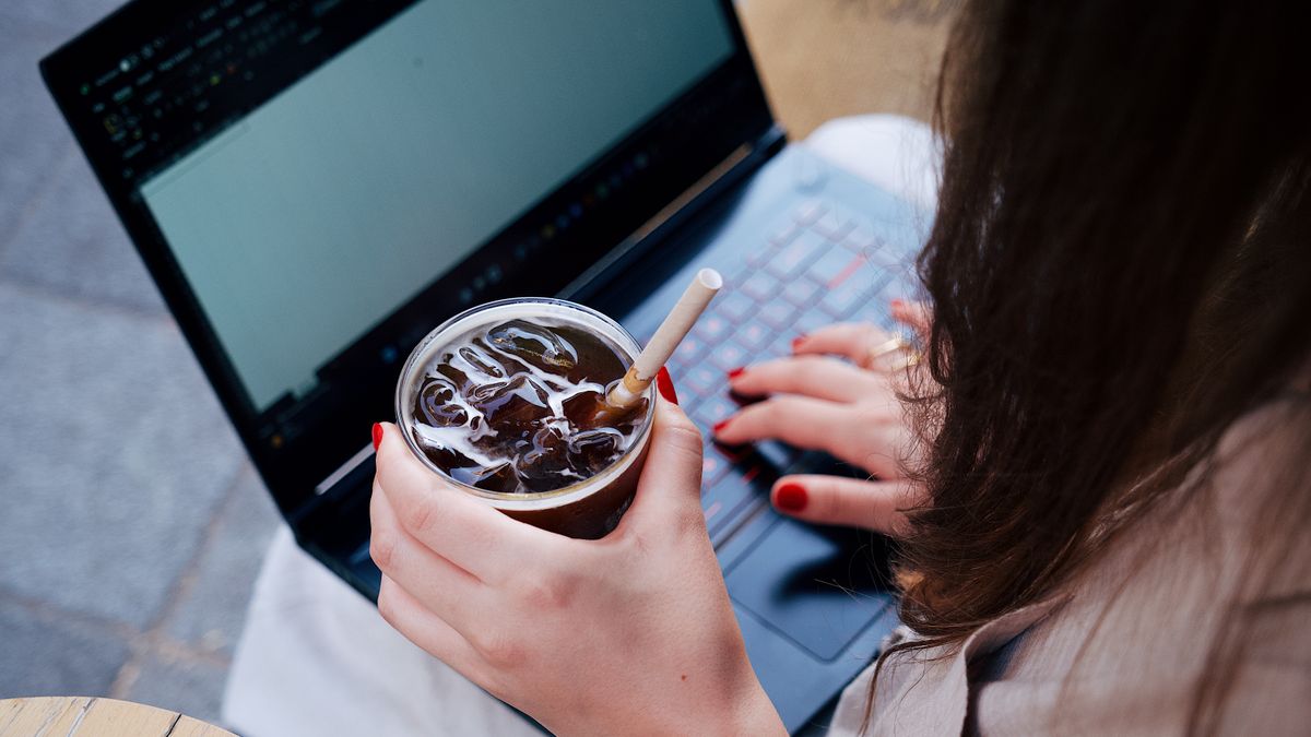An adult woman working on a laptop while holding an iced drink, seated outside