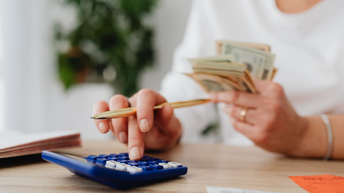 A person sits at a desk calculating finances using a calculator and holding cash
