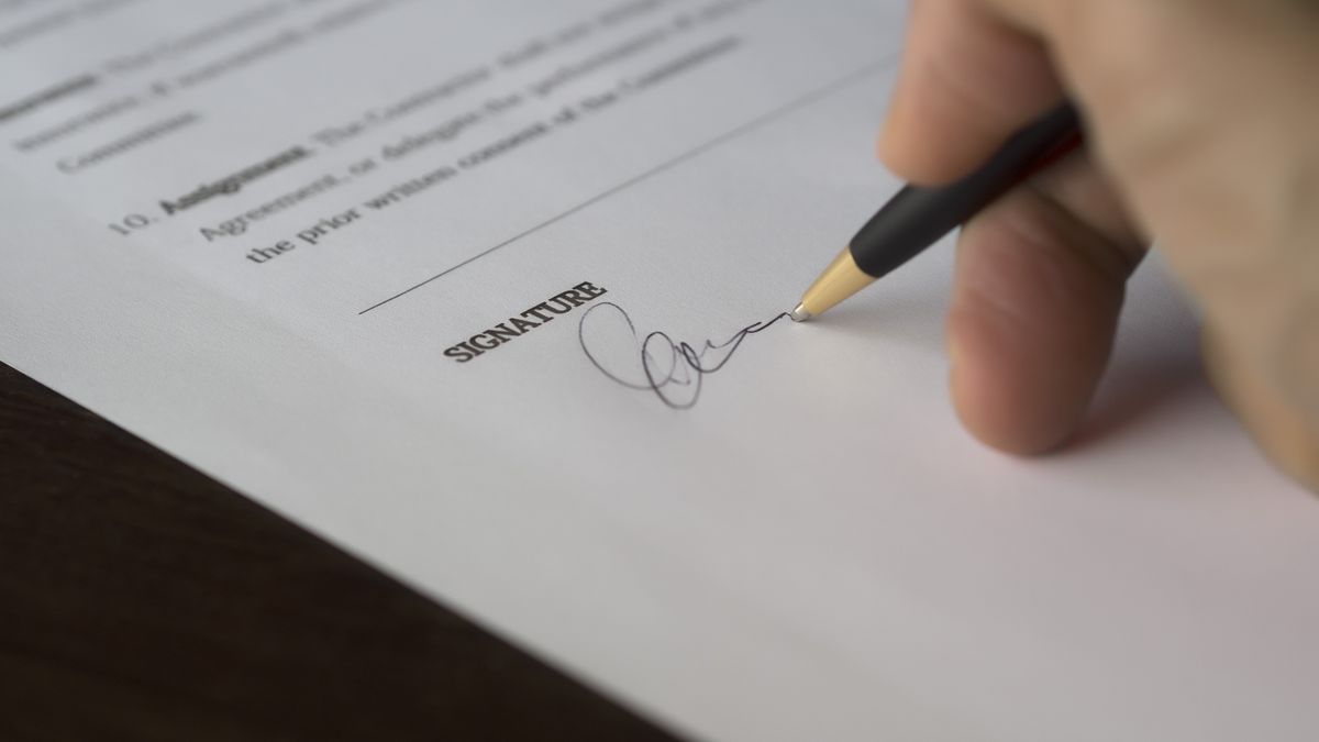 A hand signs a formal contract with a pen on a wooden desk