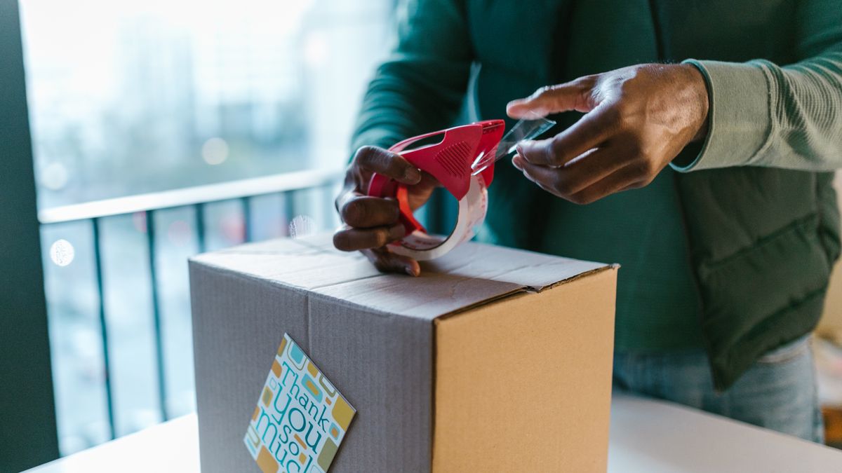 Close-up of man sealing a cardboard box with a tape dispenser indoors
