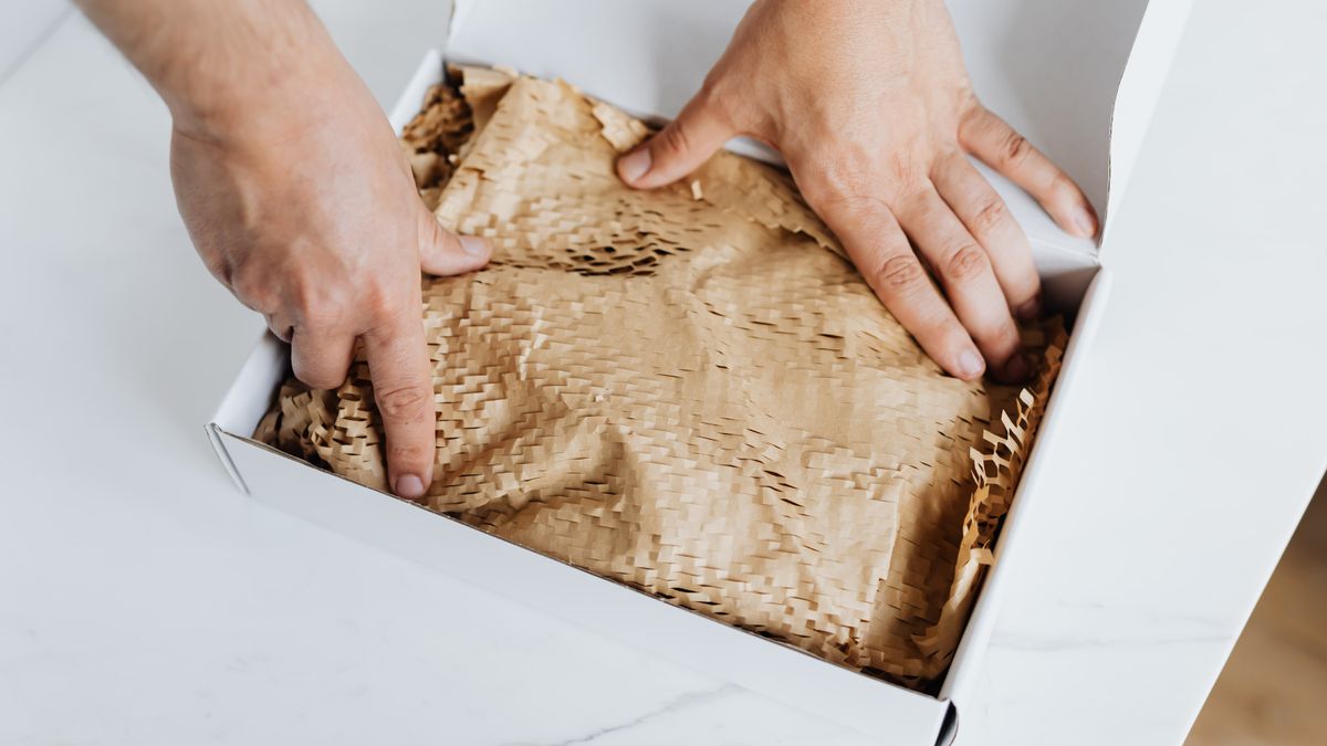 From above crop adult male packing ordered product or gift in white cardboard box with craft paper for safety postal delivery at white marble table