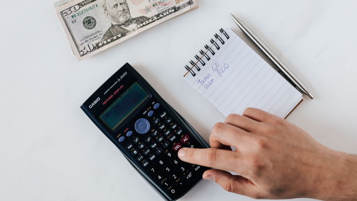 Top view of crop unrecognizable economist calculating on calculator on table with notepad and pen near pile of dollar banknotes on marble table