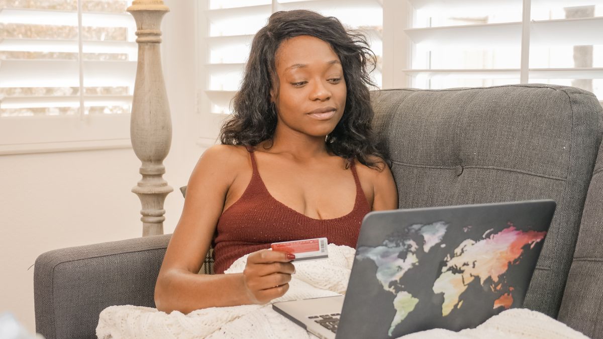 Young woman using her credit card for an online purchase while sitting on a couch with a laptop at home
