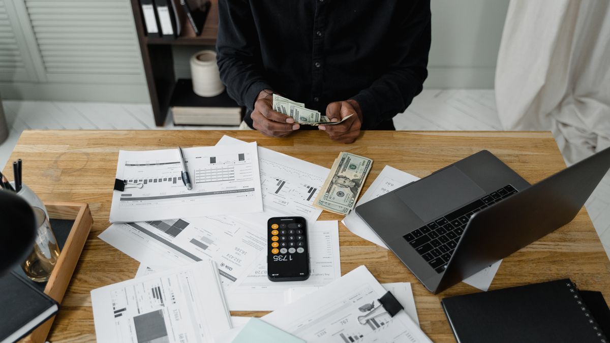 A person counts dollar bills at a desk with a laptop, papers, and calculator