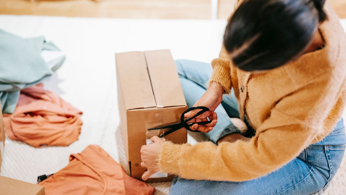 Crop anonymous female using scissors while packing carton box with parcel on floor among casual clothes